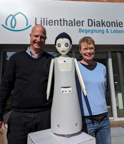 Eine Frau und ein Mann stehen vor einem Haus mit der Aufschrift "Lilienthaler Diakonie". Zwischen den beiden steht ein Roboter mit Gesicht und Mütze.