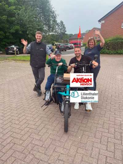 Vier Menschen. Zwei sitzen auf einem Paralleltandem, einem Dreirad. Die anderen stehen daneben. Alle Menschen winken in die Kamera.