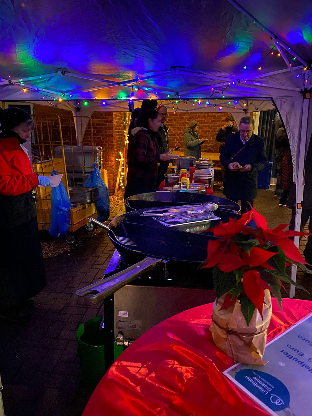 In der Dämmerung stehen Menschen draußen an der Luft in warmer Kleidung. Lichter geben ein weihnachtliche Stimmung. Auf einem Tisch steht ein Christstern. 