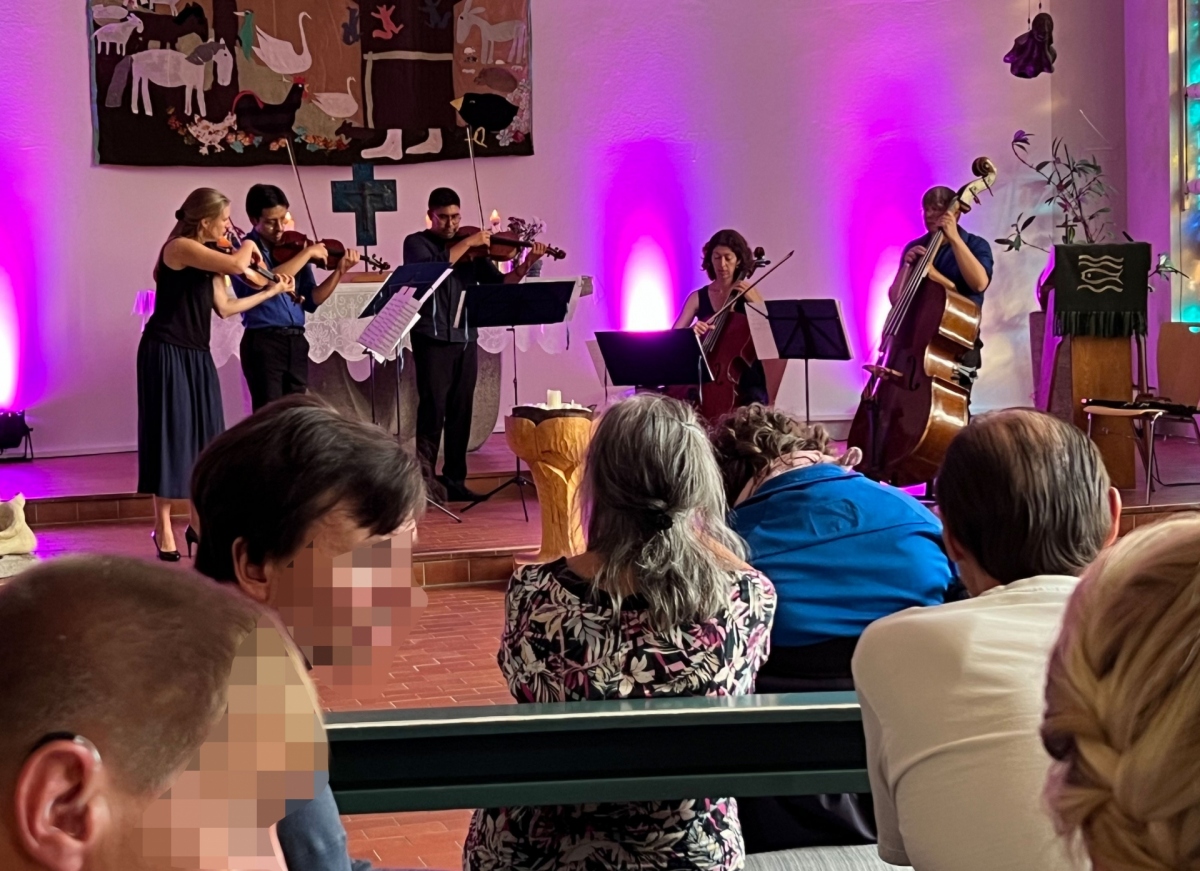 In einer Kirche stehen Menschen, die Streichinstrumente spielen. Es sitzen zuhörende Menschen in den Kirchenbänken.