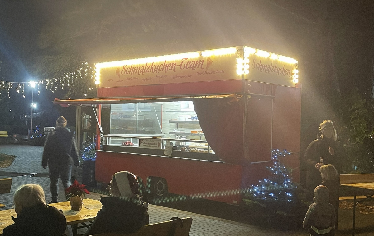 Ein Schmalzkuchenwagen steht im Dunklen, eine Lichterkette leuchtet, Menschen drumherum essen.