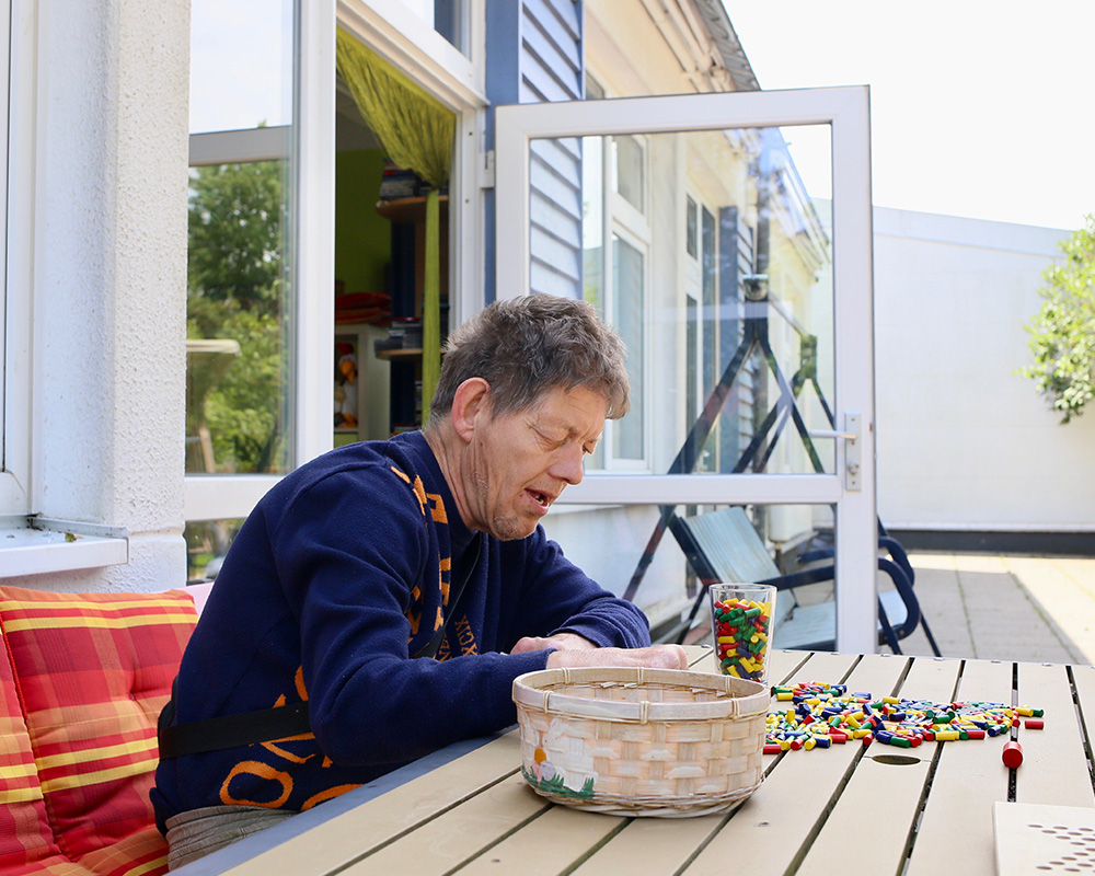 Ein Bewohner sitzt draussen vor der Tagesstätte an einem Tisch und bastelt mit Spielsteinen.