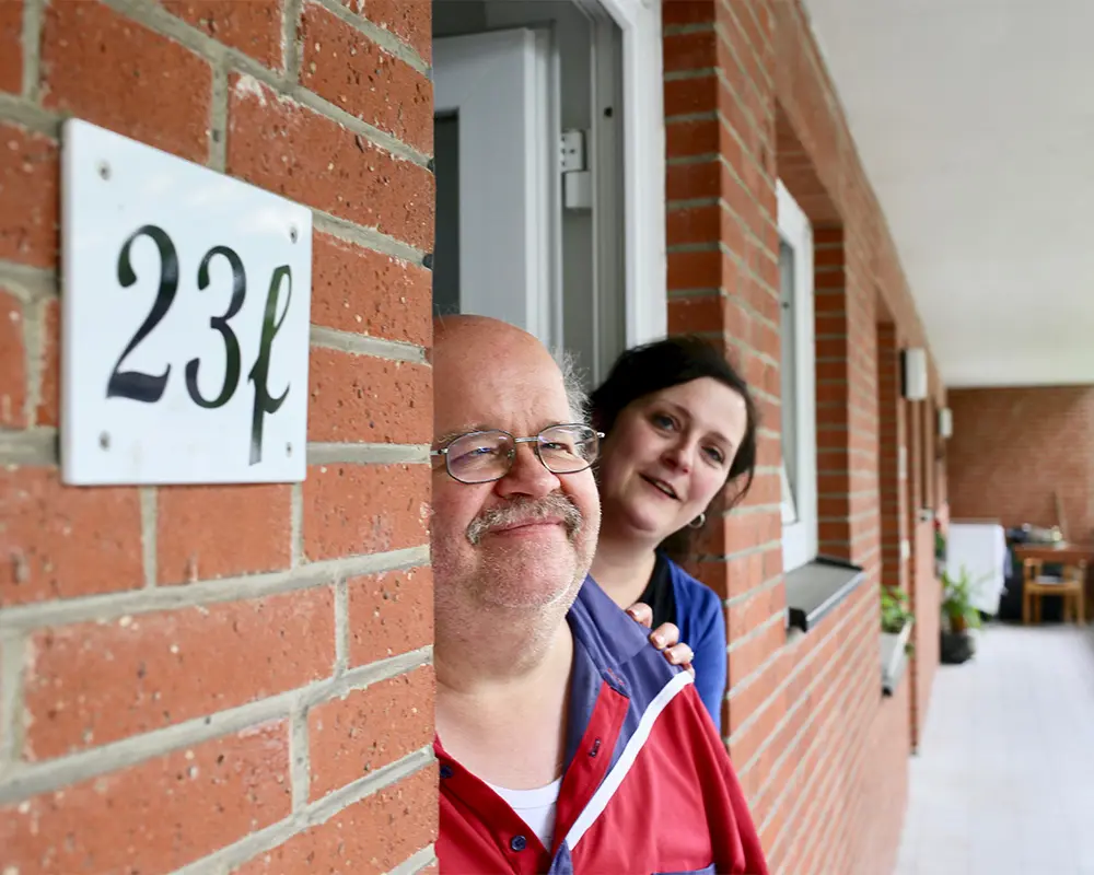 Ein älterer Herr und seine Betreuerin stehen in der Eingangstür der Wohnung und gucken freudig in die Kamera