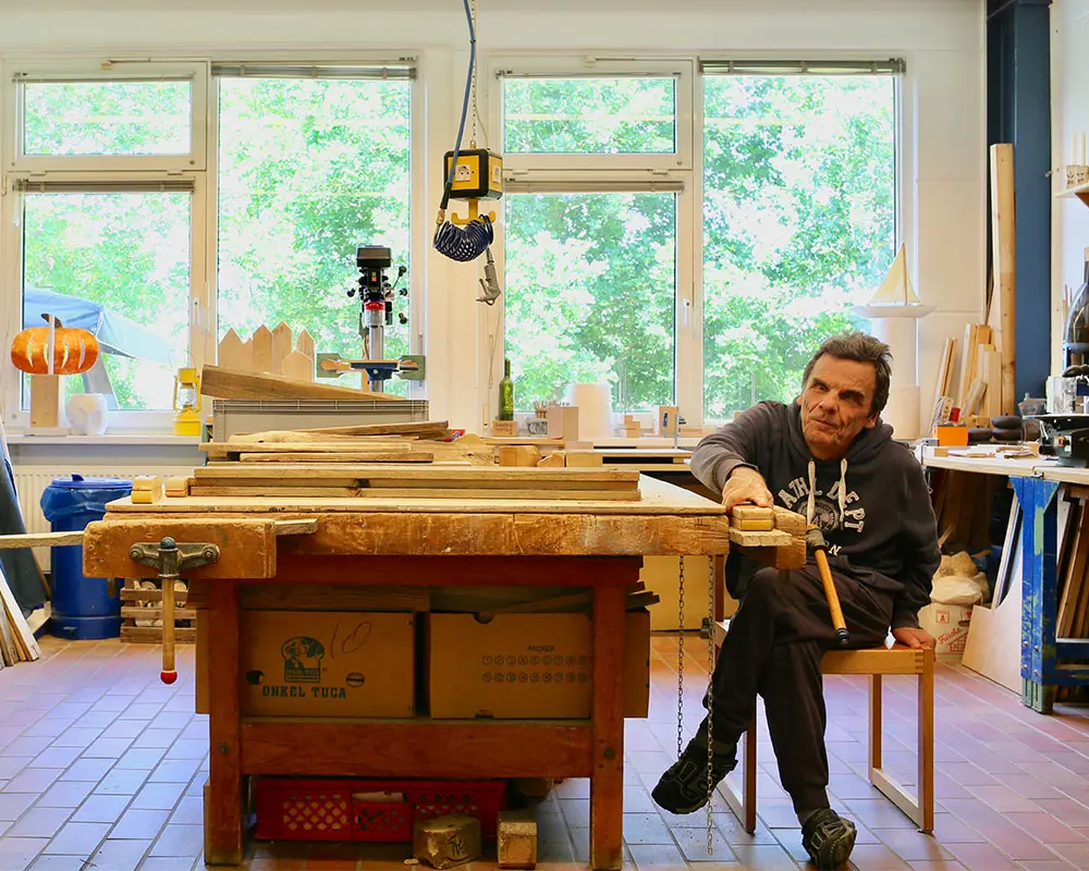 Das Bild zeigt eine Person sitzend an einem großen Arbeitstisch in einer Holzwerkstatt. Auf dem Tisch liegen verschiedene Holzlatten und Werkzeuge.