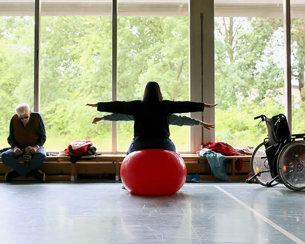zwei Frauen, sichtbar von hinten sitzen in einer Turnhalle auf einm Gymnastikball mit seitlich ausgetreckten Armen.