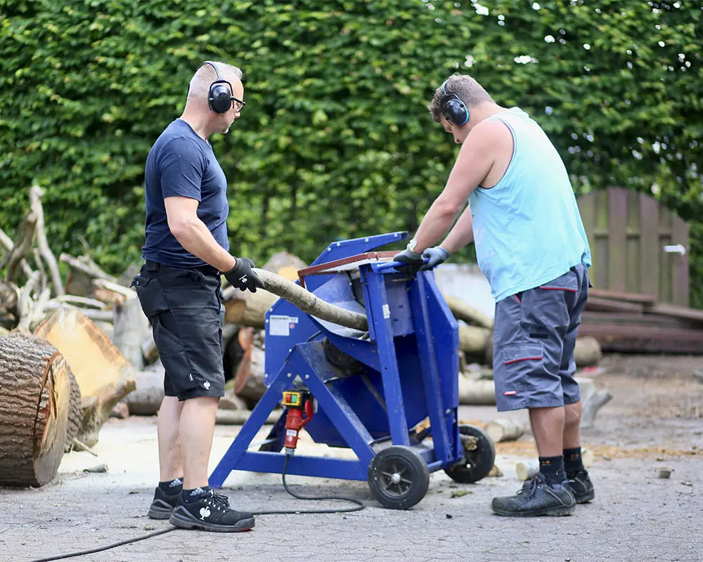 Zwei Personen stehen an einer blauen Brennholzsäge und bearbeiten gemeinsam einen dicken Holzstamm. Beide tragen Gehörschutz, Handschuhe und feste Schuhe.