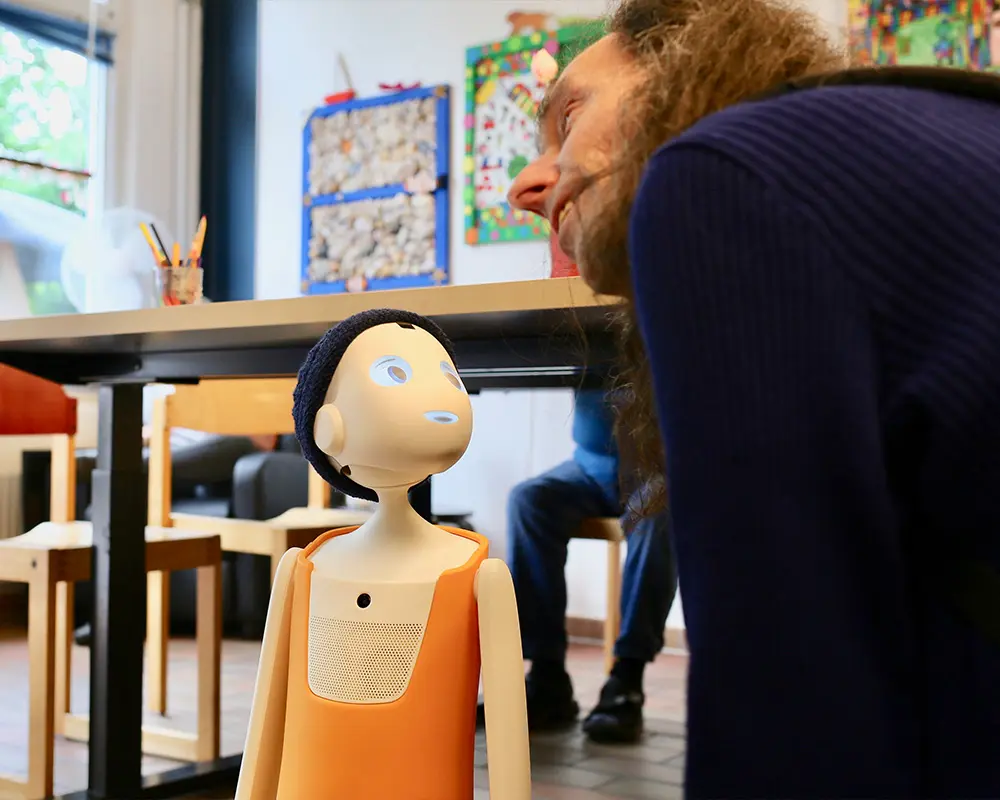 In einer Tagesstätte steht eine kleine humanoide Roboterfigur auf dem Boden. Eine Person beugt sich nah zu dem Roboter hinunter.