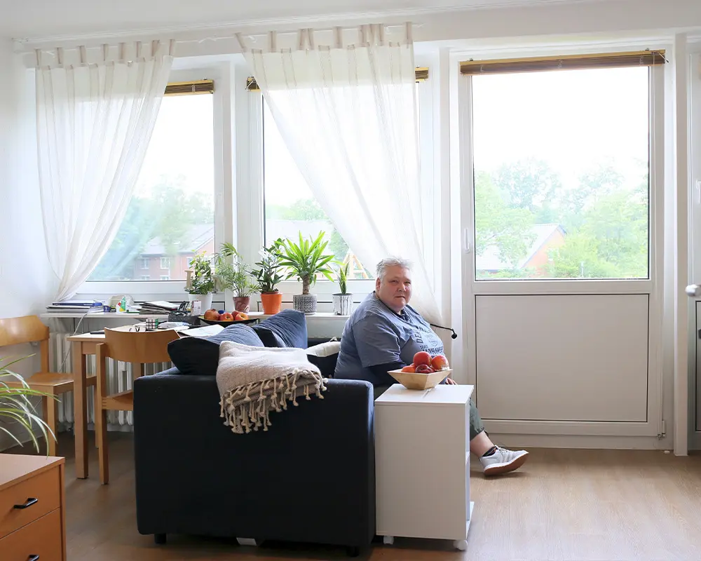 Eine Frau sitzt in ihrem Wohnzimmer auf dem Sofa. Hinter dem Sofa steht ein Tisch mit Stühlen und Pflanzen auf der Fensterbank.