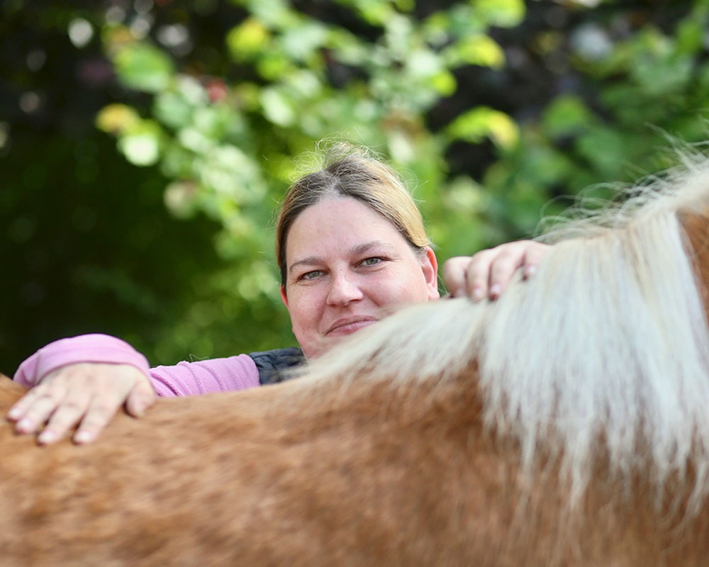 eine Fiau mit einem rosa Pullover steht hinter einem hellbraunen Pferd und hat die Hände auf den Pferderücken gelegt