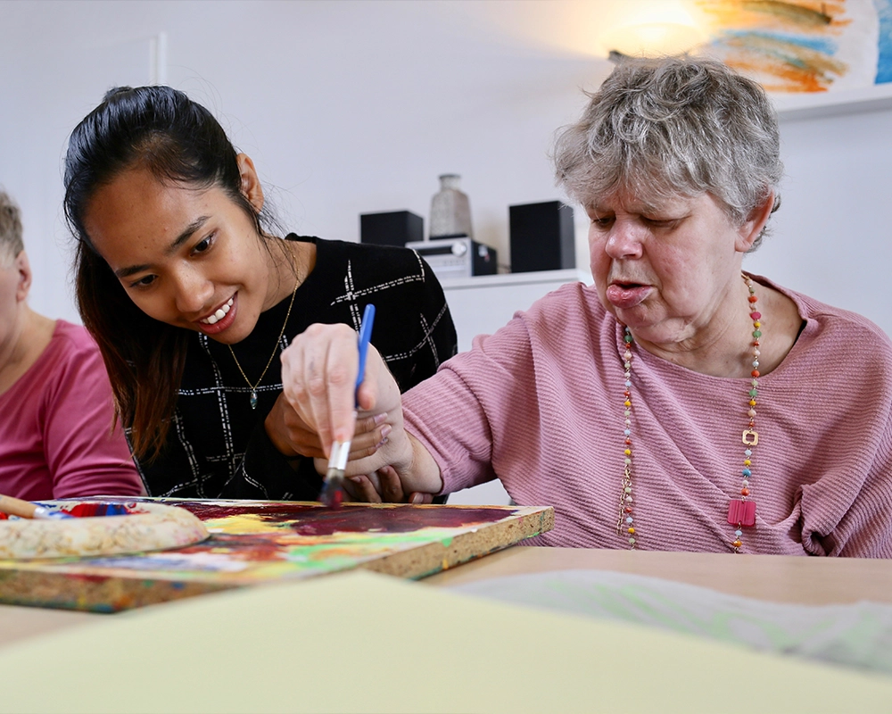 Eine Betreuerin bückt sich über ein Bild, welches von einer Bewohnering mit einem Pinsel gemalt wird.