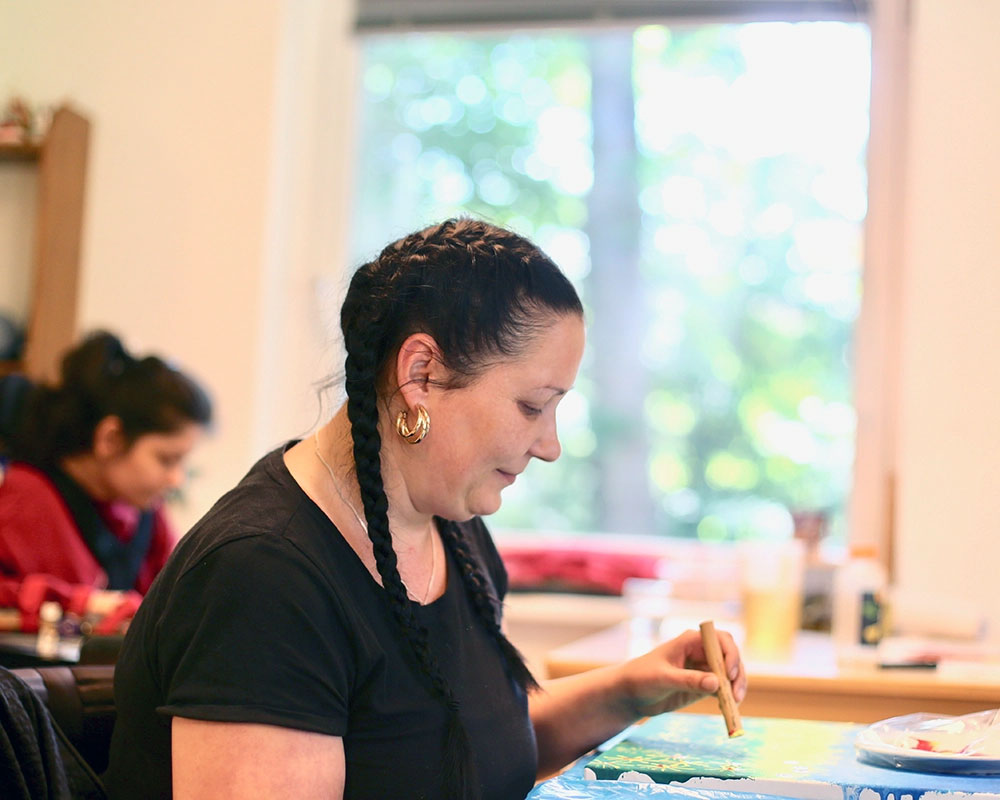 eine dunkelhaarige Frau mit einem gepflochtenem Zopf sitzt an einem Tisch und malt mit Wachstiften ein Bild