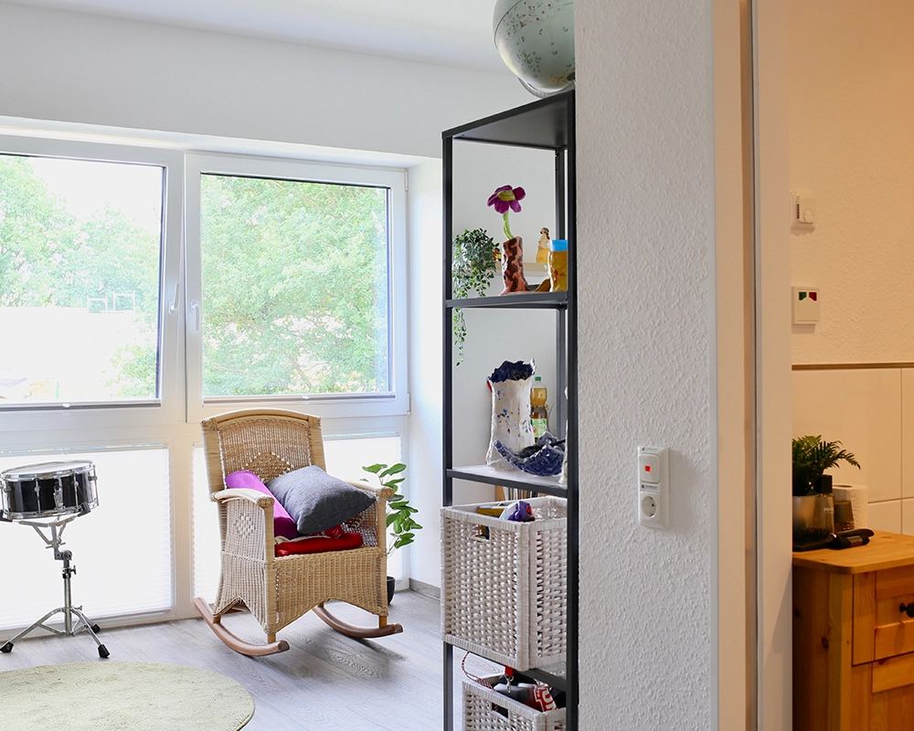 Ein heller Raum mit großen Fenstern zeigt einen gemütlichen Bereich mit einem Schaukelstuhl, einem kleinen Regal mit Deko‑ und Alltagsgegenständen.